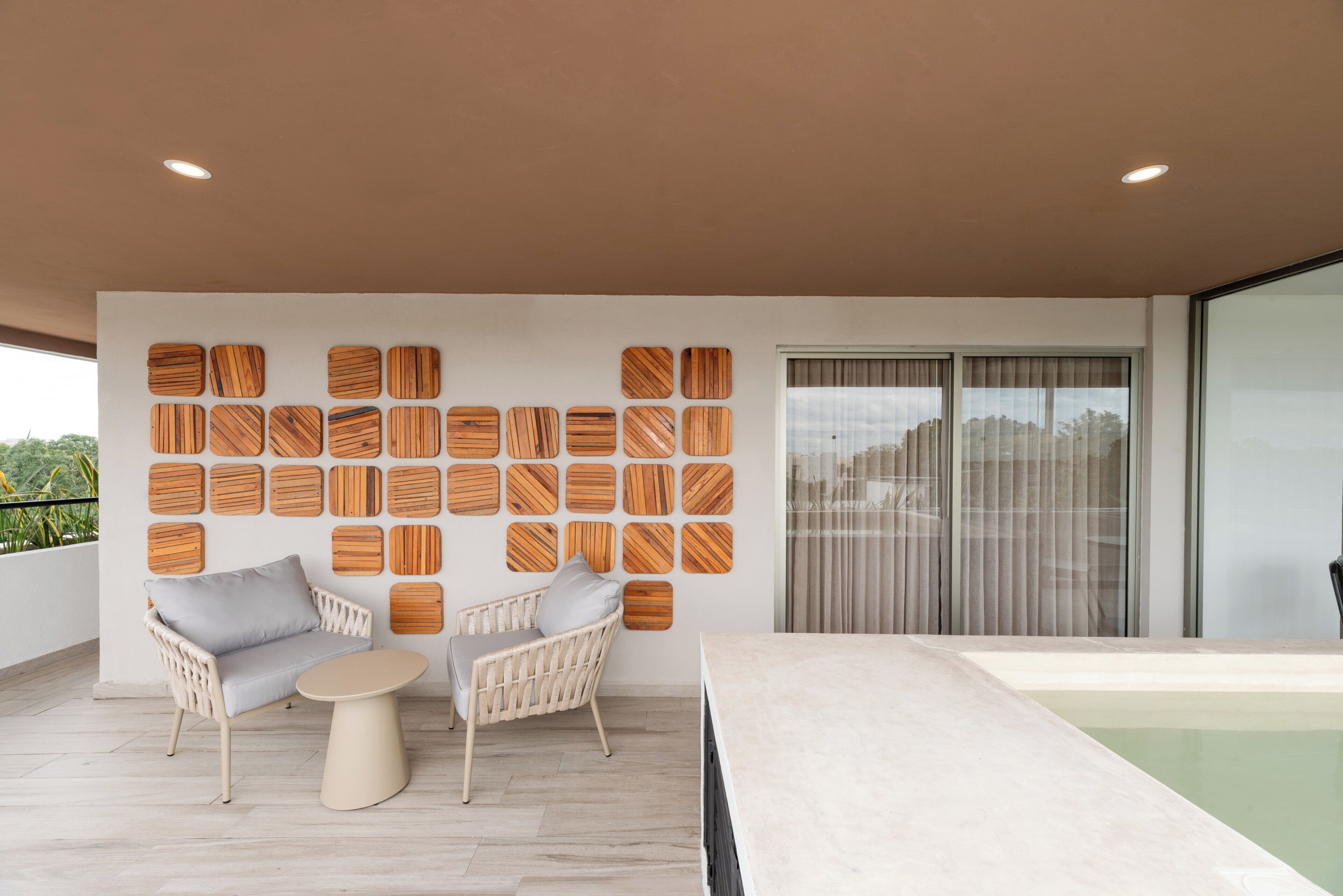 A modern outdoor seating area with two wicker chairs and a round table, a wooden wall art installation, and a large sliding glass door leading to a pool.