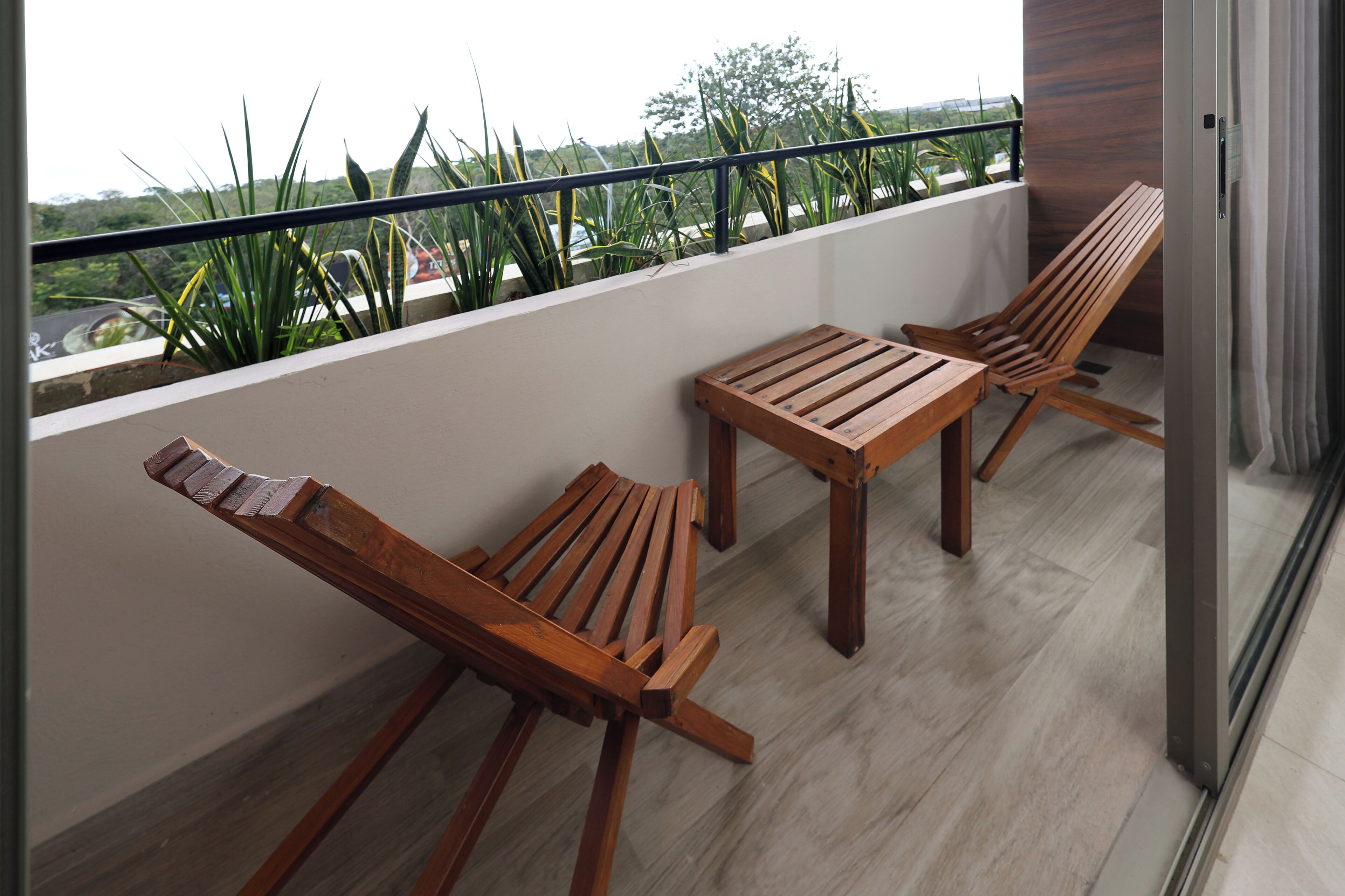 A balcony with two wooden chairs and a table, a railing, and a view of greenery and a road.