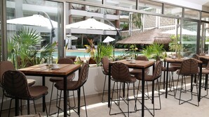 A modern outdoor dining area with tables and chairs, large umbrellas, and a view of a swimming pool and building in the background. - Holiday Inn Tulum by IHG (Tulum)