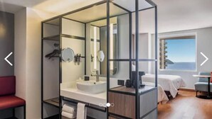 A modern hotel room with a glass-enclosed bathroom, a bed with a view, and a small table. - Fiesta Inn Mazatlán (Mazatlán)