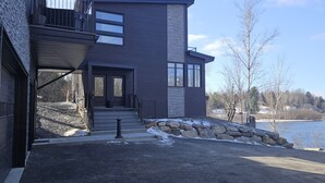 A modern two-story house with a stone foundation, large windows, and a view of a lake.