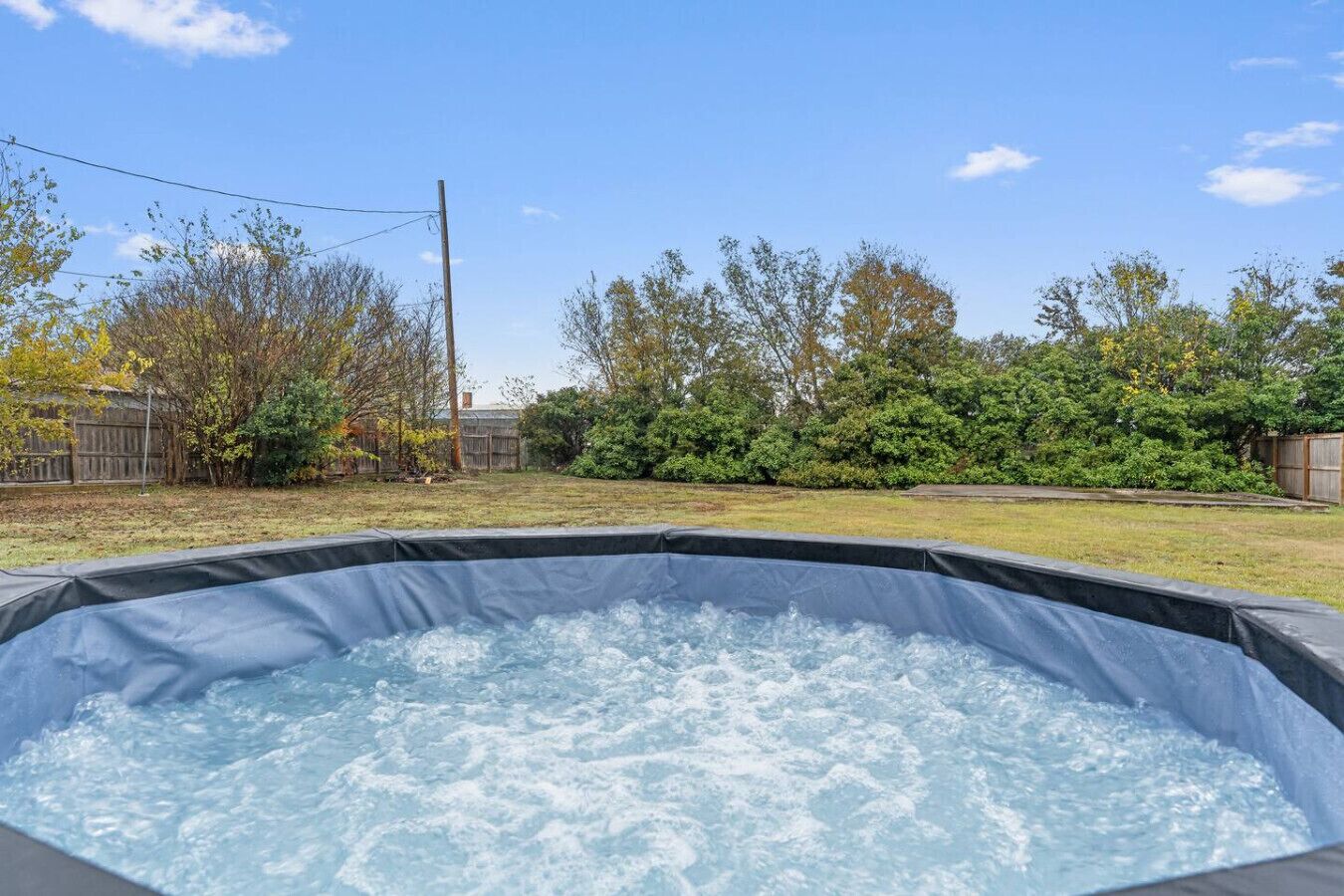 A hot tub in a backyard with a wooden fence, trees, and a clear sky.