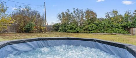 A hot tub in a backyard with a wooden fence, trees, and a clear sky.