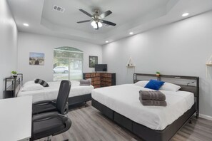 A modern hotel room with two beds, a desk, a chair, a ceiling fan, and a window with a view of a car and greenery.