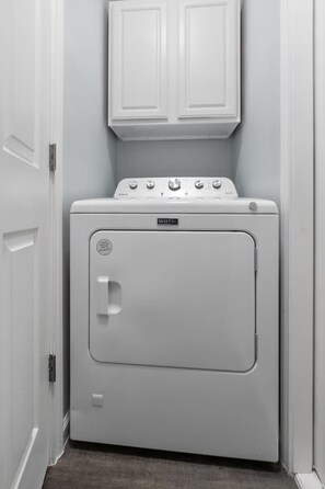 Uma máquina de lavar e secar empilhada, compacta e de cor branca, numa passagem estreita com portas e armários brancos.