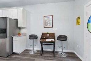 A kitchen with white cabinets, a stainless steel refrigerator, a wooden table with two bar stools, and a framed picture on the wall.