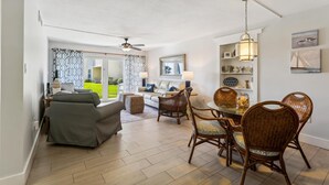 A spacious living area with a sofa, dining table, and chairs, a ceiling fan, and a view of a green garden through a sliding door. - Coquina A101 (St. Augustine)