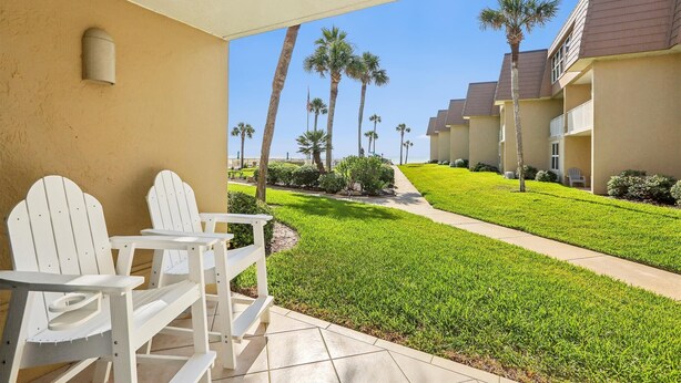 A covered outdoor area with white Adirondack chairs, a well-maintained lawn, and a view of a building with multiple balconies and palm trees. - Coquina A101 (St. Augustine)