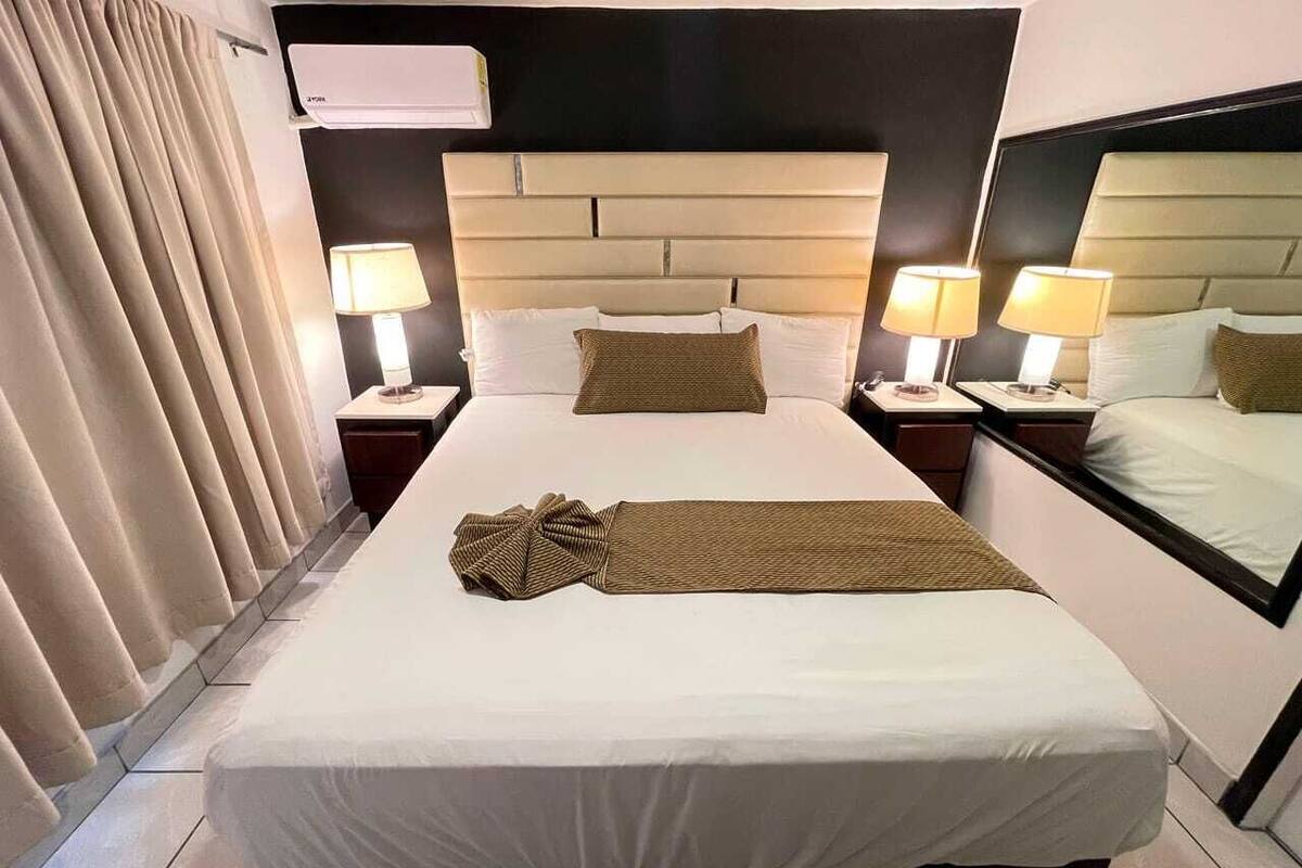 A hotel room with a large bed, two bedside tables with lamps, a headboard, and a window with curtains.