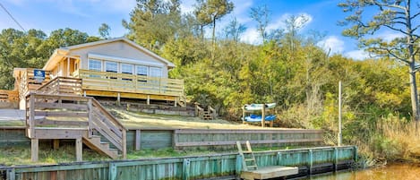 A wooden house with a deck and a dock extending into a calm body of water, surrounded by trees.