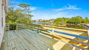Une terrasse en bois dotée d’une balustrade, qui offre une vue sur une étendue d’eau, et qui est entourée de verdure et de bâtiments situés au loin.