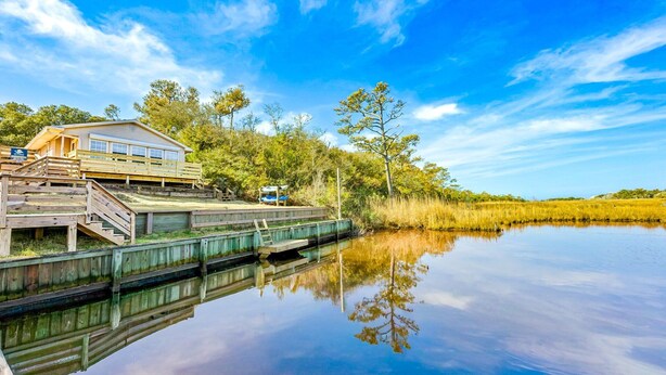Une maison en bois dotée d’une terrasse et d’un petit quai pour bateaux, entourée d’arbres et bordée par une étendue d’eau calme.