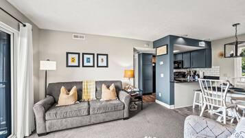 A modern living room with a grey sofa, a dining area with white chairs, and a kitchen with dark cabinets.