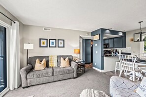 Un salon moderne avec un canapé gris, un coin repas avec des chaises blanches et une cuisine avec des meubles sombres.