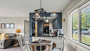 Een moderne woonkamer met een grijze bank, een eettafel met stoelen, en een zichtbare keuken door een open deur.