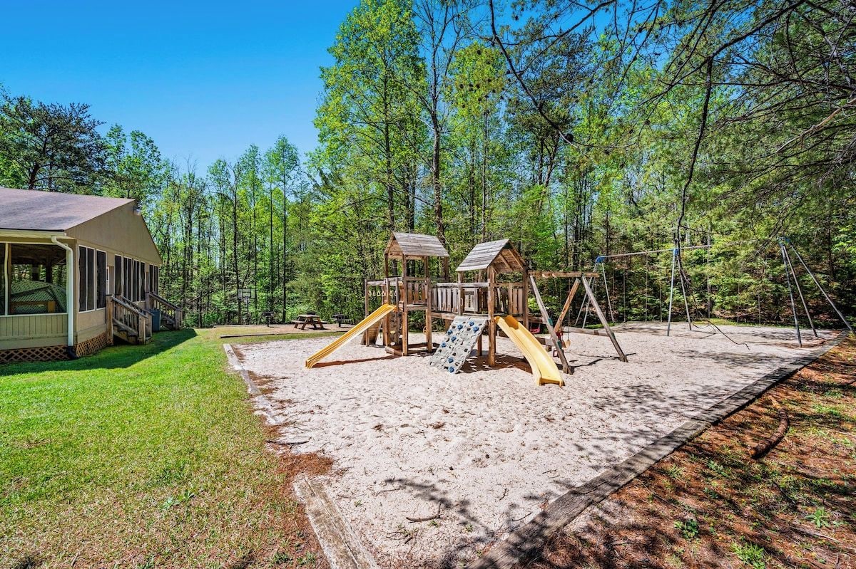 A playground with a slide and climbing structures, surrounded by a forested area.