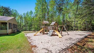 A playground with a slide and climbing structures, surrounded by a forested area.