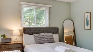 Cama bem arrumada com lençóis brancos e toalhas dobradas, mesa de cabeceira com luminária e rádio, janela com persianas e uma obra de arte com folha emoldurada na parede.