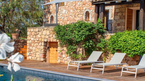 Un edificio de piedra con piscina, sillas de descanso blancas y una terraza de madera.