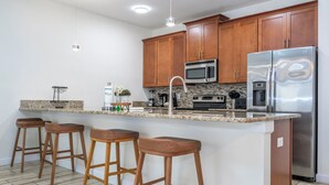 A kitchen with wooden cabinets, a stainless steel refrigerator, a microwave, and a granite countertop. There are four wooden bar stools and a sink with a faucet.