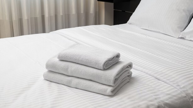 A neatly made bed with white linens and a folded towel on top, a white pillow, and a black bedside table.