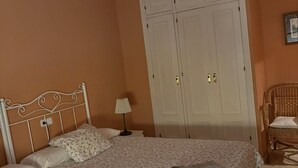 A bedroom with a double bed, a nightstand, a lamp, a wardrobe, and a wicker chair.