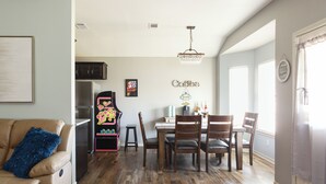 A dining area with a wooden table and chairs, a chandelier, and a kitchen in the background.