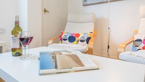 Ein Zimmer mit einem weißen Tisch, einer Weinflasche und einem Weinglas. Auf einem Stuhl liegt ein farbenfrohes Kissen und auf dem Tisch liegt ein Buch.