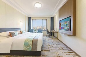Quarto de hotel com uma cama grande, uma escrivaninha com cadeira, televisão e vista da cidade através de uma janela ampla.