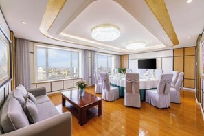 Ruime hotelkamer met een groot raam, een bank, een eettafel met stoelen en een flatscreen televisie.