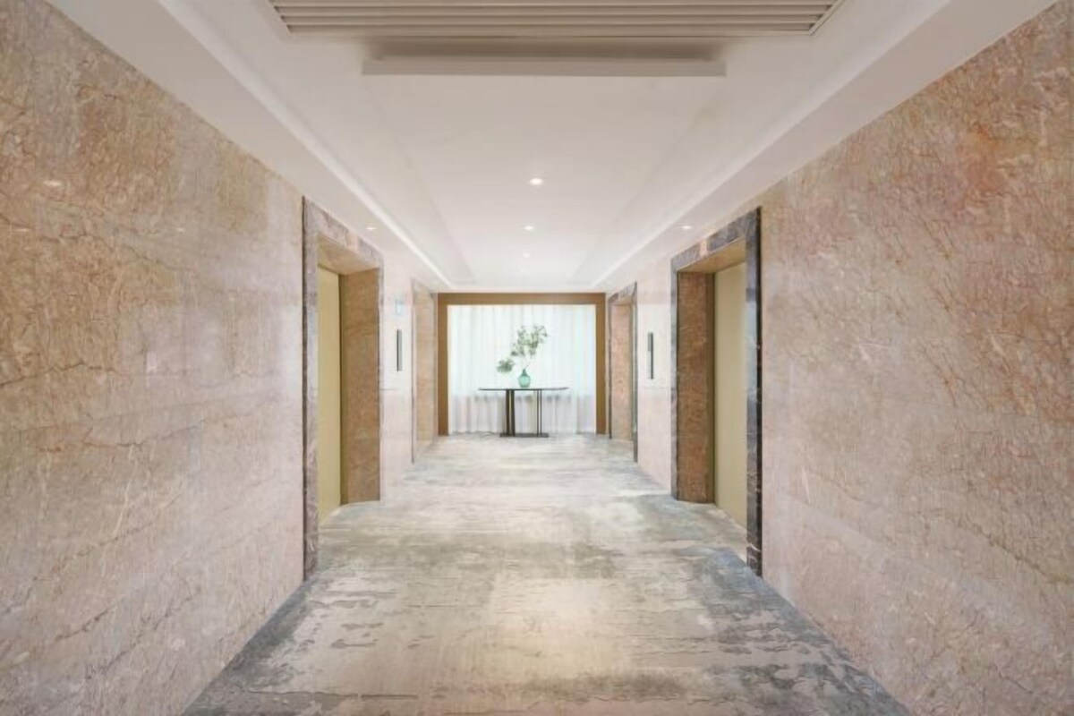 A hallway with marble walls and a concrete floor, leading to a room with a window and a table with a vase.