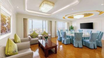 A spacious living room with a sofa, coffee table, and chairs, a dining area with round tables and chairs, and a large window offering a city view.
