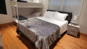 Une chambre avec un lit superposé, un lit simple, une table de chevet avec une lampe et une fenêtre avec des stores.