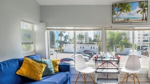 Sala de estar moderna com um sofá azul, cadeiras brancas e um carrinho. Janelas amplas oferecem vista da cidade e palmeiras.