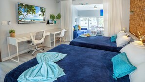 Quarto de hotel com duas camas, uma escrivaninha, uma cadeira, televisão e vista para a praia.