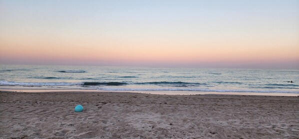 A beach with a clear sky, waves, and a single blue ball on the sand.