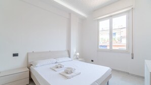 Une chambre d’hôtel minimaliste avec un grand lit, une table de chevet, une commode et une fenêtre donnant sur des immeubles.