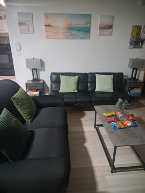 A living room with a black sofa, green cushions, a coffee table with snacks, and a painting of a beach scene on the wall. - Cozy Pocono Retreat w/ Popcorn/TVs/Pooltable/BBQ Grill/Firpit/Games/Amenities/
 (Tobyhanna)