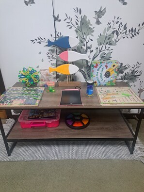 Une table en bois sur laquelle on trouve divers jouets et jeux pour enfants, une tablette et une décoration murale représentant un poisson.