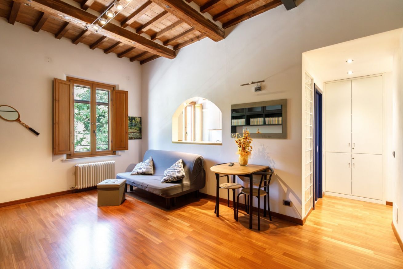 A spacious room with a wooden floor, a sofa, a small table, a radiator, a bookshelf, and a window with a view of greenery.