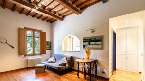 Una habitación amplia con suelo de madera, un sofá, una mesita, un radiador, una estantería y una ventana con vistas a zonas verdes.