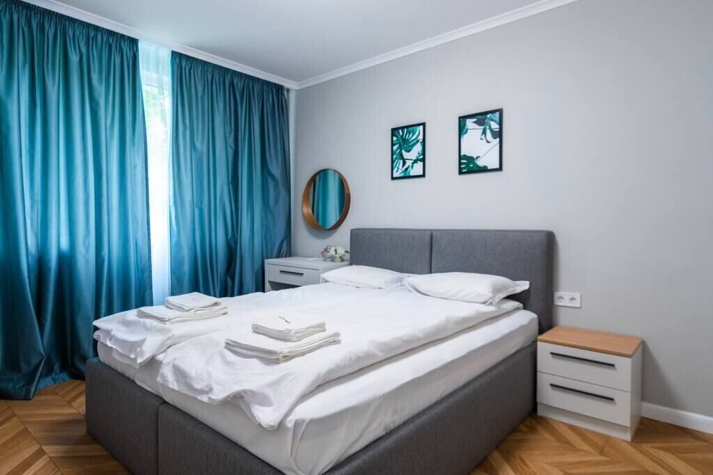 Un lit bien fait, avec du linge de lit et des serviettes blanches, une tête de lit rembourrée de couleur grise, une table de chevet avec une lampe et une petite table de nuit en bois.