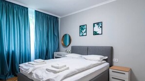 A neatly made bed with white linens and towels, a grey upholstered headboard, a bedside table with a lamp, and a small wooden nightstand. - Lozenets apartment (Sofia)