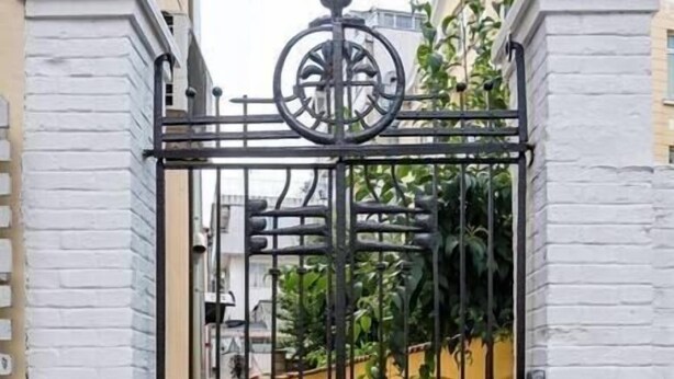 Une entrée fermée par un portail en métal décoratif, des murs en briques blanches et un chemin menant à un bâtiment agrémenté de verdure.