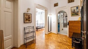 Un couloir avec un plancher en bois, des murs blancs et un miroir sur le côté gauche. On aperçoit un lit à travers une porte ouverte au fond.