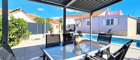 A covered outdoor seating area with a table and chairs, a pool in the background, and a clear view of the surrounding buildings.