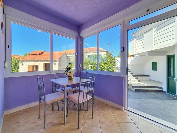 A room with a dining table and chairs, a vase with flowers, and large windows with a view of buildings and trees.