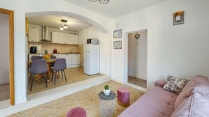 Una sala de estar moderna con un sofá rosa, una mesita redonda con una planta y un comedor con sillas, con una cocina al fondo.