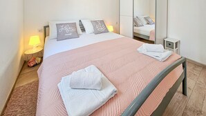 Una cama bien hecha con un edredón rosa y dos toallas blancas dobladas, una lámpara de noche y una mesita de noche con lámpara.