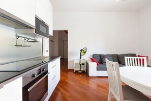 Una cocina moderna con horno empotrado, una zona de comedor con mesa y sillas blancas, y un sofá gris con cojines rojos.